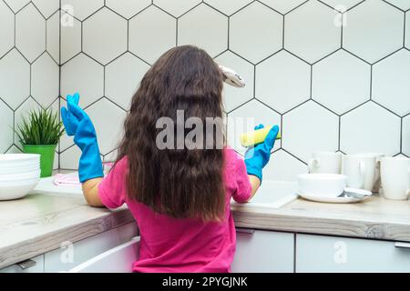 Little girl in household gloves washing up tableware with dishwashing sponge and dish soap in kitchen sink. Clean home. Stock Photo
