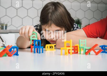 Thoughtful, focused little girl building and holding small green chair toy near many colorful chair toys on white table Stock Photo