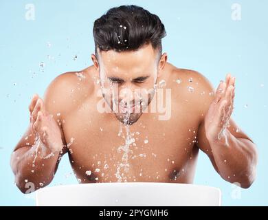 Face, water splash and skincare of man cleaning in studio isolated on a blue background. Hygiene, water drops and male model washing, bathing or grooming for healthy skin, facial wellness or beauty. Stock Photo