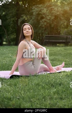A young positive woman in a gymnastic suit practices yoga and meditates while sitting on a mat. Stock Photo