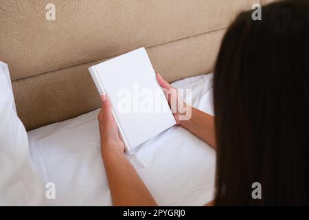 Young woman holding blank notebook or book mockup with white cover while lying on the bed. Stock Photo