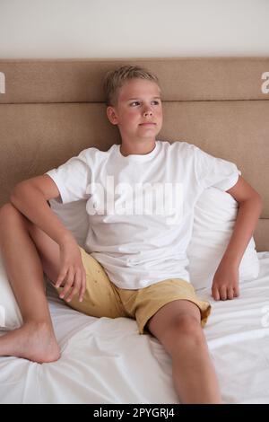 A cute young teenager sits in bed in a white t-shirt and looks out the window. Stock Photo