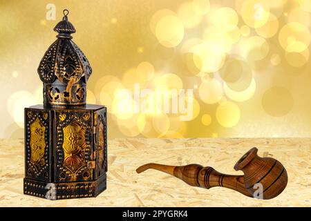 Old oriental or arabic metallic lantern and a handmade wooden tobacco pipe on table over abstract golden stars background. Ramadan Kareem template. Card design. Stock Photo