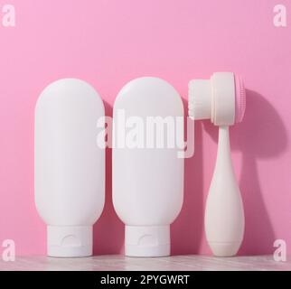 White plastic tubes with cream and a massaging brush for facial cleansing on a pink background, items for cosmetic procedures Stock Photo