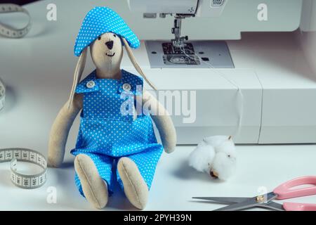 Funny smiling sewed stuffed rabbit toy in blue overalls, cocked hat, sit near cotton, scissors, next to sewing machine Stock Photo