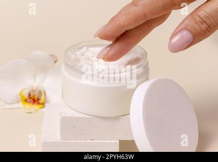 A woman hand using cream from Opened jar with a blank lid near orchid flowers, mockup Stock Photo