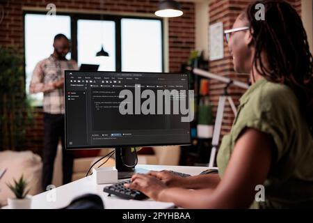 Selective focus on computer screen with programming language text of programmer working remote in home living room. African american coder developing code on pc while boyfriend is using laptop. Stock Photo