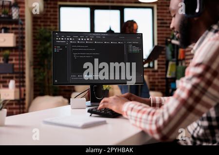 Freelance programmer with wireless headphones writing code on personal computer while girlfriend is using laptop standing in living room. African american man developing software debugging algorithm. Stock Photo
