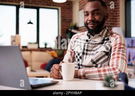 African american programmer posing confident looking at camera holding a cup of coffee while girlfriend is reading a book. Freelancer coder taking a break relaxing watching video content on laptop. Stock Photo