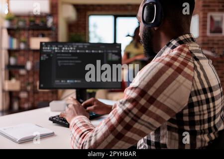 African american programmer with wireless headphones listening to music while writing code on personal computer. Freelance coder developing software while enjoying online radio on headset. Stock Photo