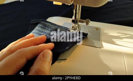 Sewing process on a modern sewing machine, close-up. Sewing machine foot with a needle. Dark blue fabric is trimmed with double wrapping of the fabric edge. Natural sunlight Stock Photo