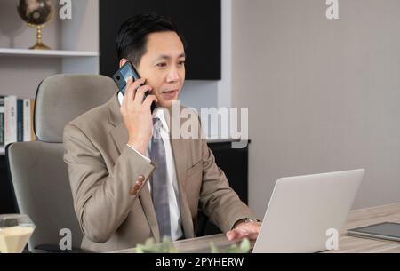 Businessman talking on smartphone and working on laptop computer at office desk. Stock Photo