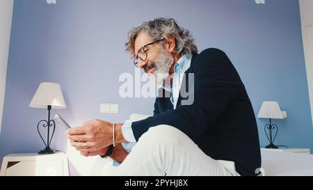 Stylish good looking mature man is sitting on bed and using smartphone Stock Photo