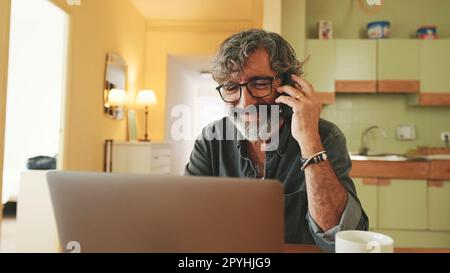 Mature businessman working on laptop, talking on mobile phone sitting in home kitchen Stock Photo