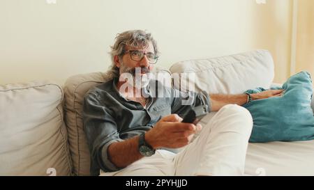 Close-up of the hand of an elderly man with remote control, changes channels while sitting on the couch at home Stock Photo