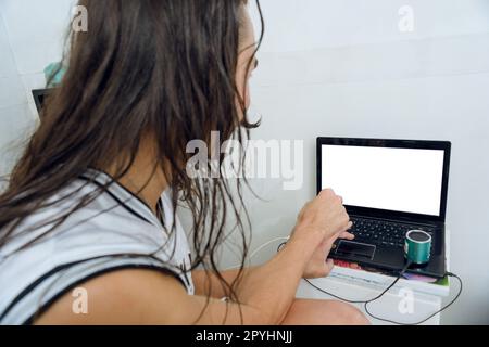 rear view of young gender non-binary person with long hair in bedroom sitting on bed looking at white laptop screen, copy space Stock Photo