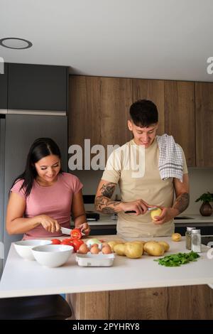 Two young latin people peeling potatoes and cutting tomatoes at kitchen. Stock Photo