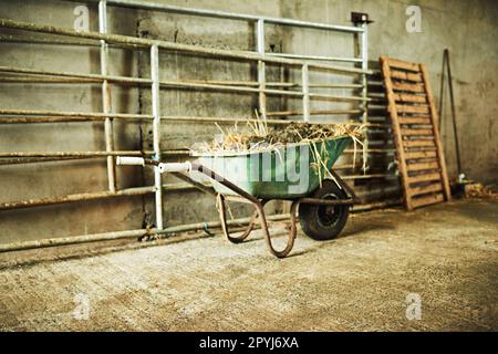 Use a wheelbarrow to transport things around the farm. a wheelbarrow filled with dirt and hay inside a barn on a farm. Stock Photo