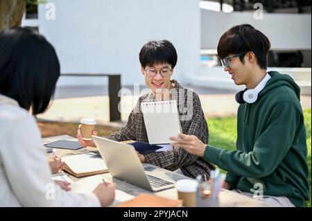 Smart young Asian male college student is tutoring math to his friends at a campus park. Two Asian male college students are discussing work while wor Stock Photo
