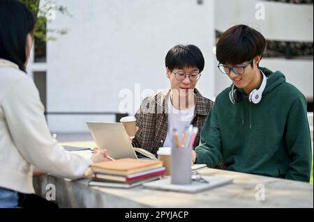 Joyful and happy young Asian male college student talking with his friend while sitting with his group at a table in a campus park. Stock Photo