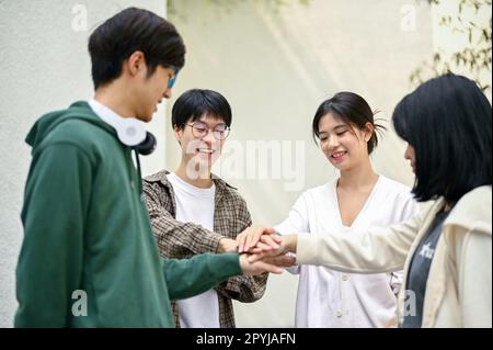 Image of a group of cheerful Asian college students putting their hands together, showing teamwork and unity. Stock Photo