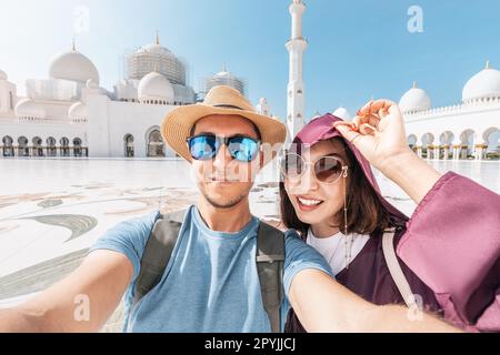A young tourist girl wearing a traditional Abaya and man in hat taking selfie photo of their honeymoon journey at Sheikh Zayed Grand Mosque in Abu Dha Stock Photo