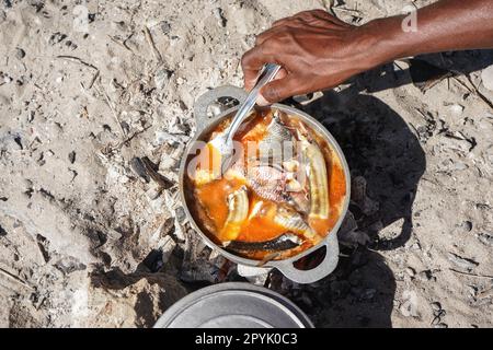 Fish soup or stew cooked from freshly catch seafood in aluminium pot, closeup detail to local man hand from above, sun shines on sand near Stock Photo