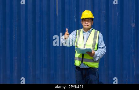 Senior contractor in safety vest and yellow hardhat holding document clipboard, stand with finger thumbs up in front of a large cargo container. Stock Photo