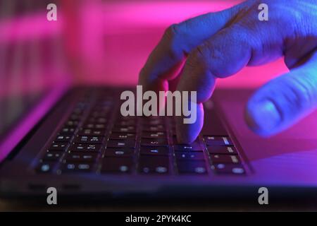 Person typing at laptop computer at night. Male uses keyboard, shakes buttons with fingers. Internet security, cyber attack. Trendy neon light. Creati Stock Photo