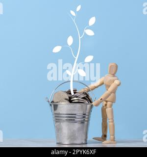 A wooden dummy and a miniature bucket with metal coins and a painted tree with leaves on a blue background. Income and profit growth concept Stock Photo