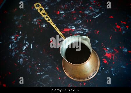 Balkan cezve for turkish coffee. A copper cezve on black surface. Antique bronze utensils. Carved metal handle with pattern. Black background with white, red and blue spots. Stock Photo