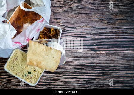Chinese takeaway food selection in plastic bag. Bami, babi pangang, crispy shredded beef, sweet and sour chicken, krupuk top view on wooden table copy space. Food delivery Stock Photo