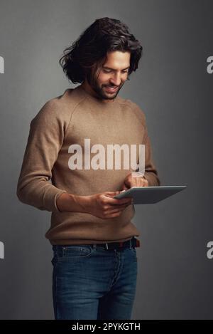 Tapped into the latest trends. Studio shot of a handsome young man using a digital tablet against a gray background. Stock Photo