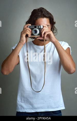 Freezing a moment in time. Studio shot of a young man taking a photo with a vintage camera against a grey background. Stock Photo