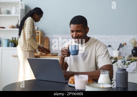 African American man using laptop while having breakfast at table in the kitchen with his wife cooking in background Stock Photo