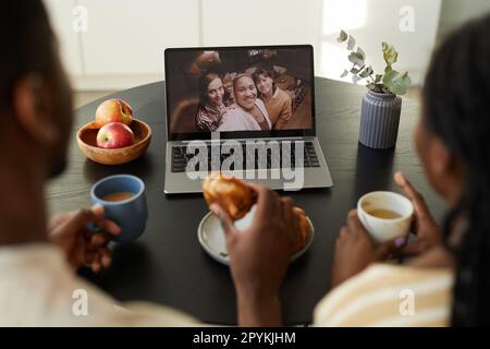 Rear view of young couple sitting at table in front of laptop and having breakfast while having video call with friends Stock Photo