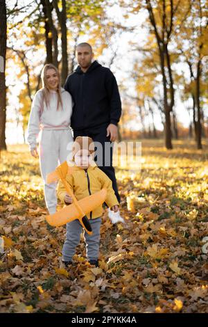 Father, mother and little son play together in autumn park or forest. Child launching orange toy airplane. Family spending good time outdoors. Stock Photo