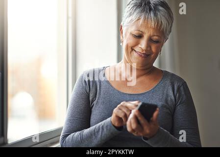 Window, typing and senior woman with a cellphone, social media and mobile app at home. Mature female, happy model and old lady with a smartphone Stock Photo