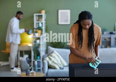 African American young woman wiping dust from furniture during housework at home together with her boyfriend Stock Photo