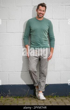 Portrait looking at camera of cheerful bearded adult man wearing casual clothes leaning against white wall. A smiling mature man poses for a photograp Stock Photo