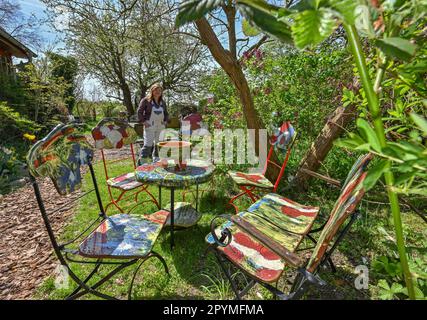 04 May 2023, Brandenburg, Sieversdorf: Undine Gomille, artist, stands next to ceramic chairs and tables and prepares the garden for the 'Open Studio Days'. The first weekend in May has become a fixed date for many art enthusiasts over the past few years. On May 06 and 07, 2023, artists will again open their studios in Brandenburg's counties and independent cities. Those who like can discover painting, graphics and book art directly at the place of origin, can look over the shoulder of sculptors and ceramic artists, learn to understand printing techniques and in some places even get the opportu Stock Photo