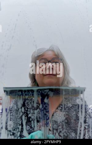 close-up of a white-haired woman cleaning soap-covered windows with a squeegee Stock Photo
