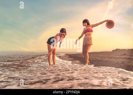 Two young Caucasian young women in their twenties, wearing shorts and bikinis, playfully splash water on their feet at the beach during sunset. One ho Stock Photo