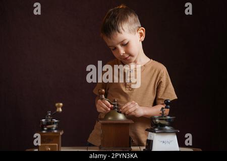 Cute preschool boy grinds coffee in a manual retro coffee grinder with interest. Studio photo on a brown background. Stock Photo