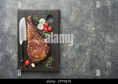 Tomahawk steak. Sliced grilled tomahawk beef steak with baked cherry tomatoes, herbs and salt on old wooden background. Preparing to grill.  Top view Stock Photo