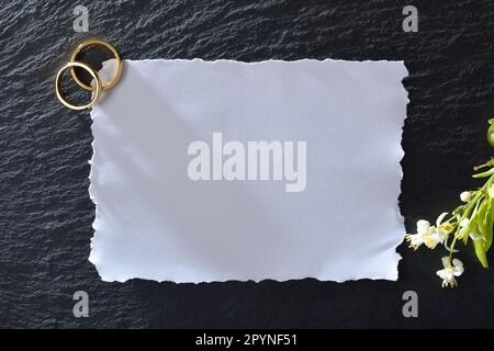 Wedding invitation with empty white paper for text on slate stone with two gold rings in one corner and flowers in another. Top view. Stock Photo
