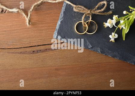 Detail of two gold rings joined with jute rope on slate stone on a rustic wooden table and flowers. Top view. Stock Photo