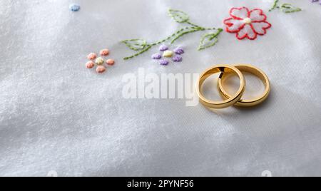 Detail of two gold wedding rings lying one on the other on white fabric with floral embroidery. Top elevated view. Stock Photo
