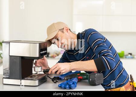 Professional young repairman repairs a coffee maker. Handsome worker in uniform repairing coffee machine in a workshop. Stock Photo