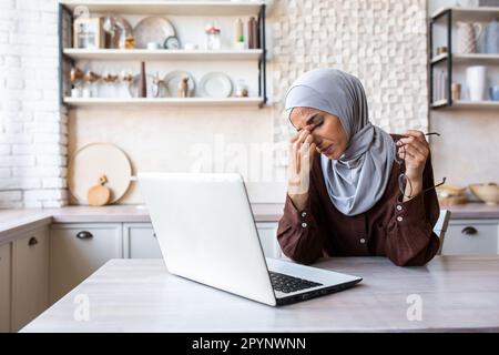 Muslim young woman sitting upset at home in the kitchen using a laptop. Read bad news, received a letter, got tired of online work. He rubs his eyes with his hand. took off and holds glasses, resting. Stock Photo